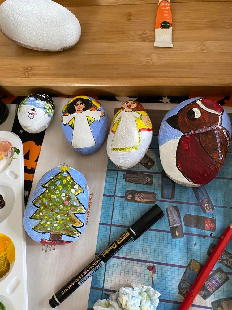 The picture is of five newly painted Christmas pebbles, with the side  of a paint palette and a black Pentel pen. One pebble has a snowy night scene with a snowman and two Christmas trees in front of mountains, two pebbles have angels, one pebble a robin and one a Christmas tree.