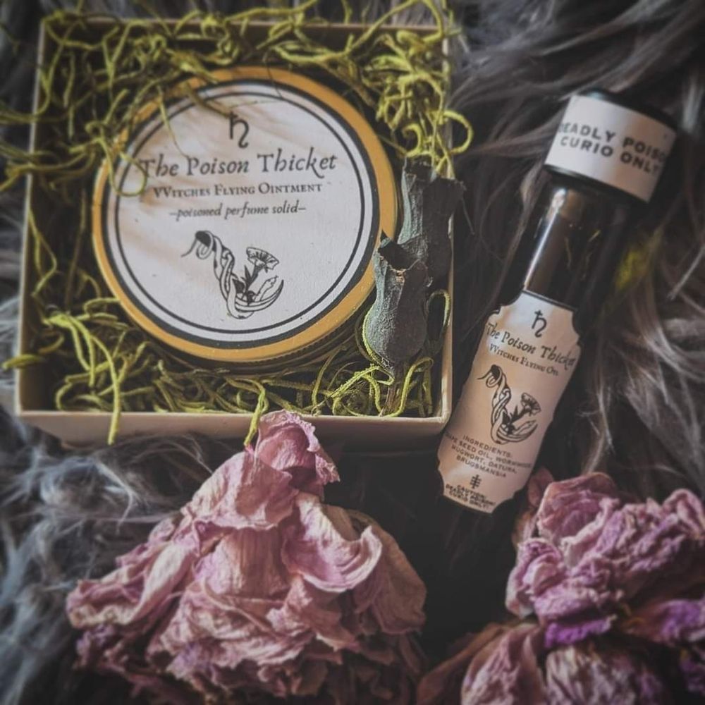 Vial of witches flying oil and tin of witches flying ointment solid perfume are staged against a light gray faux fur background. The perfume is to the upper right sitting nestled in some dried green moss and a henbane seed pod within a small beige box. The vial of oil is tucked diagonally between two dried pink peonies. Both products vintage apothecary style labels displaying the brand name The Poison Thicket, as well as product information, Saturnian planetary symbols, and a caution label that says: 'Deadly Poison, Curio Only'.
