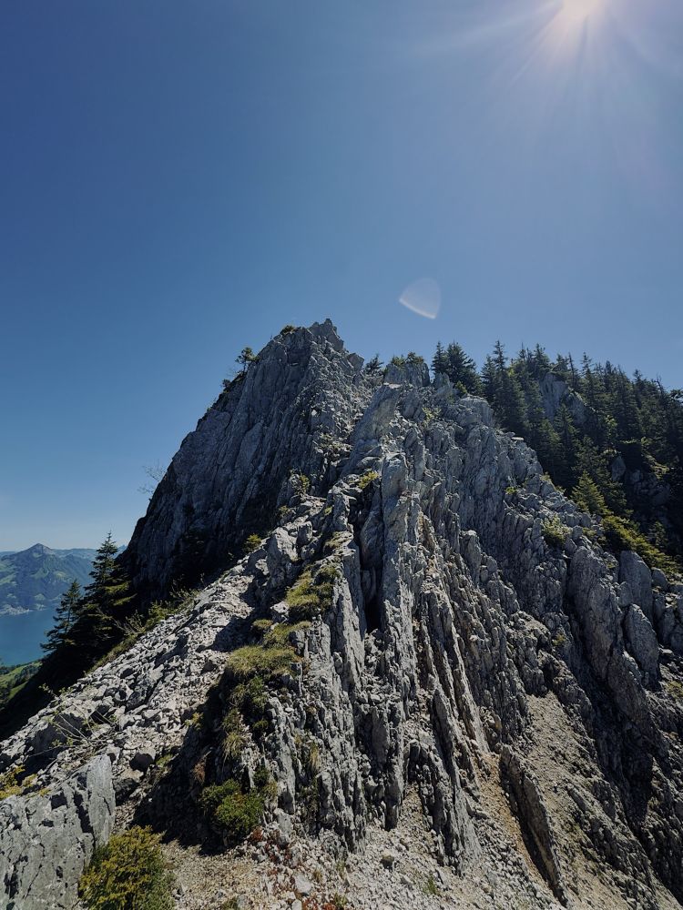 Ein schroffer, felsiger Berggrat ragt dramatisch in den blauen Himmel, von der Sonne hell beleuchtet. Der Pfad verläuft entlang der schmalen Felskante, gesäumt von wenigen Nadelbäumen. Im Hintergrund sind alpine Landschaften und ein türkisfarbener See zu erkennen – ein eindrucksvoller Ausblick auf die Bergwelt.