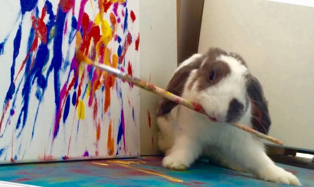 A bunny holds a paintbrush in its mouth, painting bold, colorful strokes on a canvas.