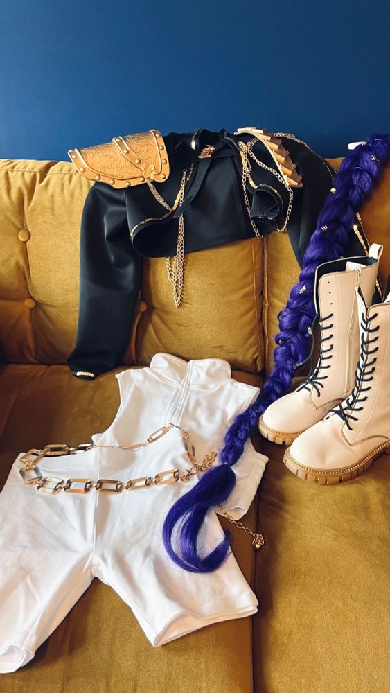 Die Einzelteile des Rumi Cosplays aus kpop demon hunters auf einer Gold gelben Couch vor einer dunkelblauen Wand. Wir sehen weiße Stiefel mit Gold Sohle, eine weiße Hose, ein weißes top, ein goldener Gürtel und eine aufwändig verzierte schwarze Jacke mit vielen Gold Elementen und einer goldenen schulterplatte. Das lila Haarteil mit dem langen Zopf liegt auch dabei 