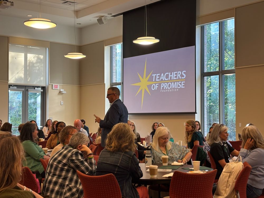 Keynote speaker Wade Whitehead addresses a group of attendees seated at round tables during the VACTE conference session. A large screen behind the speaker displays the logo for “Teachers of Promise Foundation”. The setting is a bright, modern room with tall windows and natural light. The image conveys a collaborative, professional learning environment focused on teacher development.