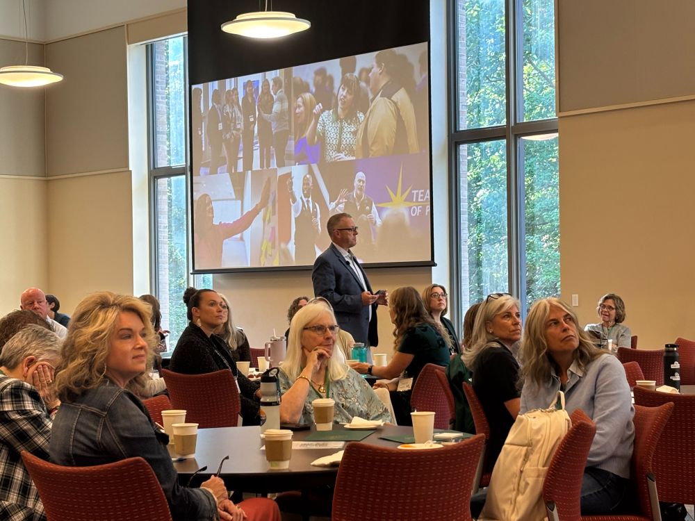 Keynote speaker Wade Whitehead stands in front of a large screen displaying a collage of images from previous educational events during the VACTE conference session. Attendees are seated at round tables in a bright room with tall windows and natural light. 