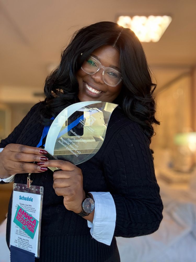 Glory Munthali holding a clear glass “Donald Matteson Outstanding Graduate Student of the Year Award". A conference badge labeled “Schedule” and “AMHCA Welcome Reception” hangs from a lanyard around Glory's neck. The background shows an indoor setting with soft lighting.