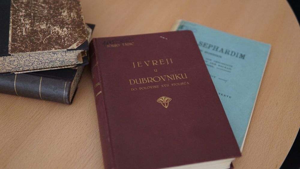 Mehrere historische Bücher auf einem Holztisch, darunter im Vordergrund ein dunkelrotes Buch mit dem Titel "Jevreji u Dubrovniku do polovine XVII stoljeća" (Juden in Dubrovnik bis zur Mitte des 17. Jahrhunderts) von Jorjo Tadić. Dahinter ist ein hellblaues Buch mit dem Titel 'Sephardim' zu sehen.