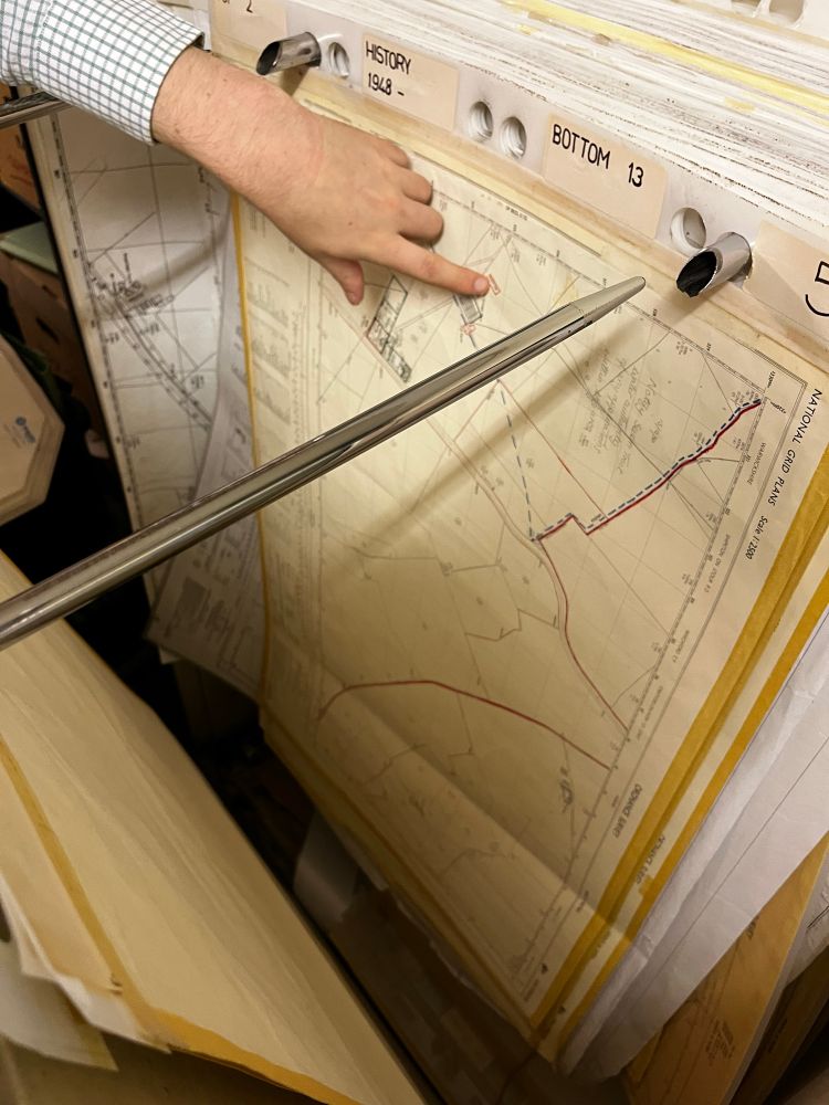 A person in a checkered shirt examines historical maps stored in a large flat filing drawer. The open drawer reveals aged survey maps or architectural plans with red boundary lines and technical drawings.