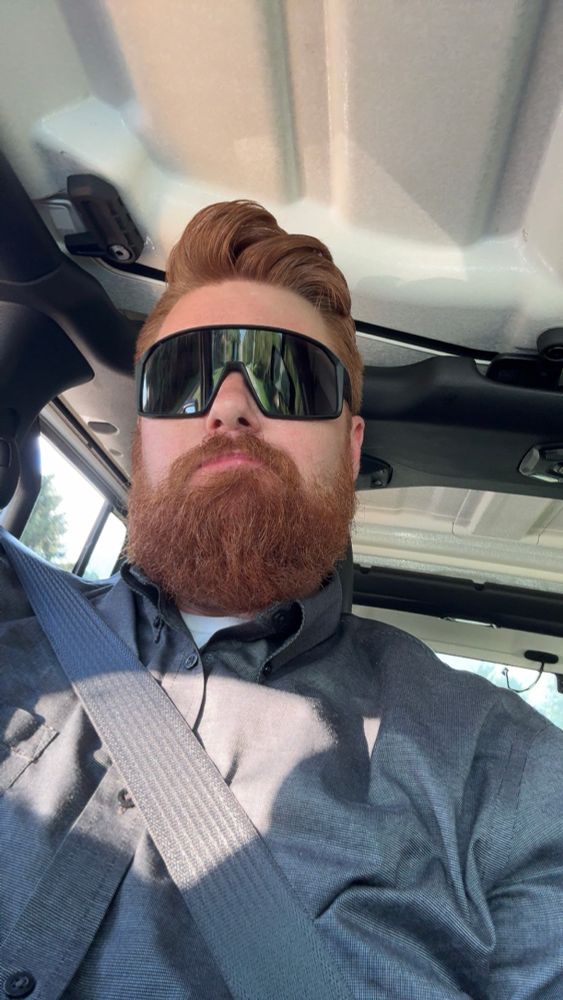 Ginger with a beard wearing douche sunglasses with a bear smile in a white jeep with his seatbelt on