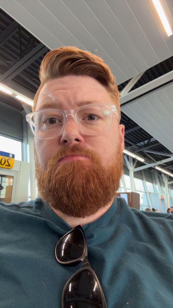 Bearded Ginger with clear white glasses with sunglasses tucked into a blue shirt, sitting at gate B20