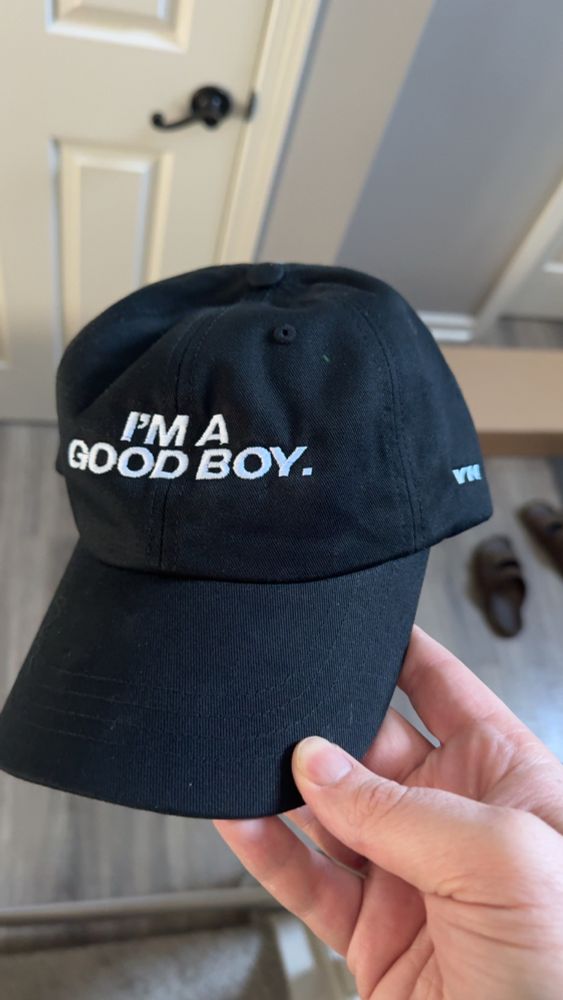 Holding a black hat with the words “I’m a good boy.” embroidered in white.