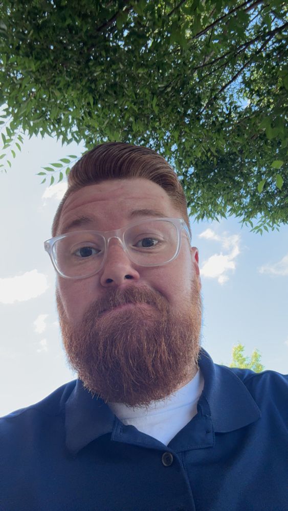 Ginger with a beard, fresh haircut with a tree and blue sky behind him.