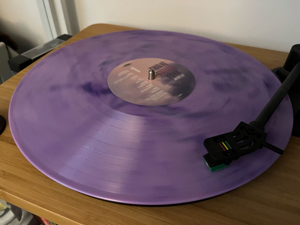 A marbled pastel purple record spinning on a turntable. The sticker in the middle depicts clouds in the sky, also tinted purple with a warm glow to the sky.