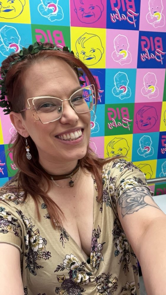 A woman with red hair and gold glasses, in a v neck beige floral blouse and a greenery crown taking a selfie in front of a neon pop art style background