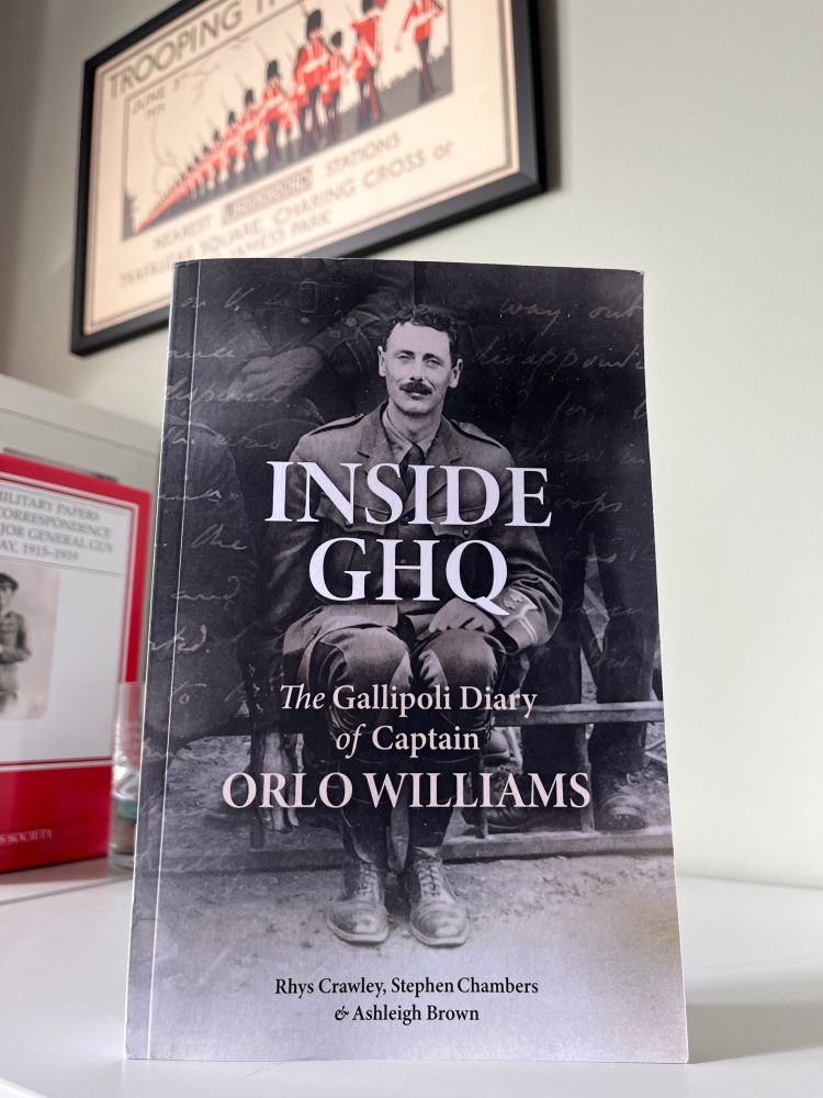 A copy of Orlo Williams' diary on a grey bookshelf. Behind the book is a poster advertising the British Army's "Trooping the Colour". Williams' diary has a picture of Orlo on the front cover in his First World War khaki uniform. Across the picture of Orlo is the title of the book, Inside GHQ.