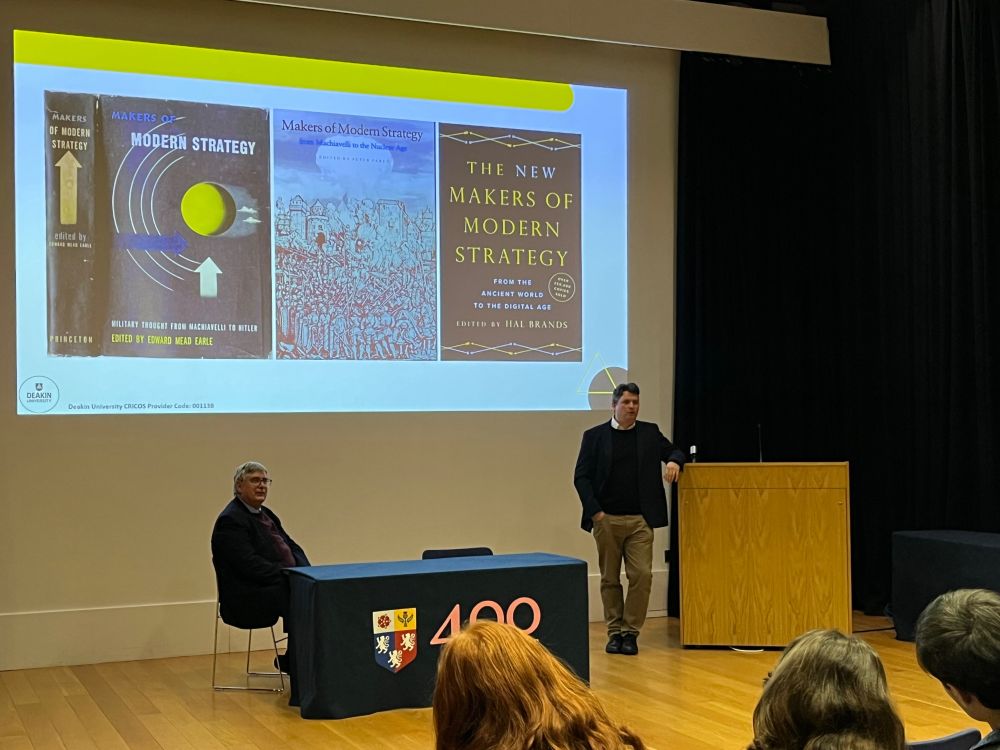 Mike finch standing at lectern in front of a slide with three covers of the three editions of Makers of Modern Strategy. Prof Adrian Gregory sits at a table to Mike’s right. 