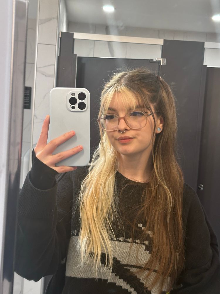 A mirror selfie of Shania in a movie theatre bathroom, she has half brown half blonde split dye hair, big round circle glasses and wearing a comfy jumper. She’s looking at the phone head tilted with a slight smile