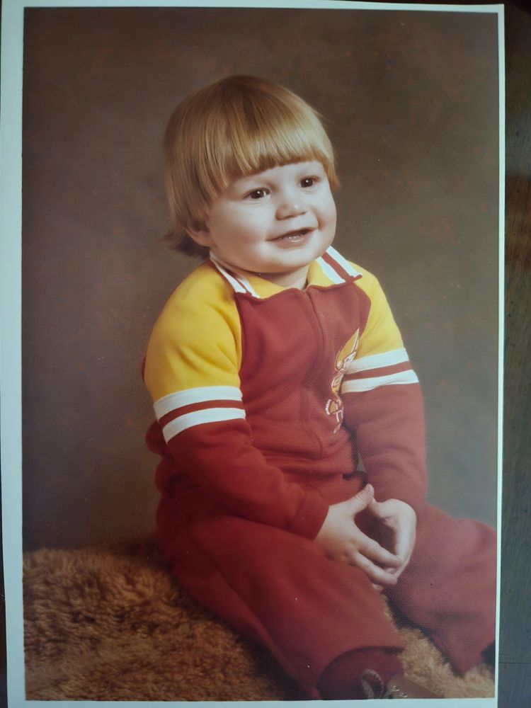 Large pale baby wearing a Washington Redskins onesie with his thumb and fingers pressed together. 