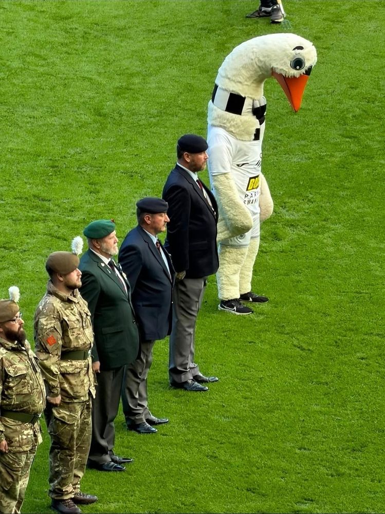 A row of military and formally dressed men observing a moment of silence. At the end of the line is a giant goose with its head bowing in respect. 