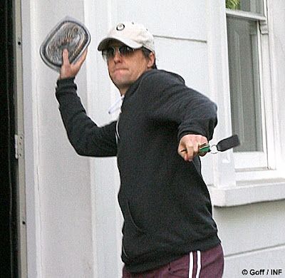 Hugh Grant wearing a dark hoodie, white baseball cap. Holding keys in one hand. With his other hand he is throwing a plastic container of baked beans directly at the camera. 
