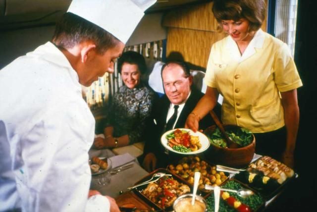 Interior of an airplane where a chef in actual chef's clothes is serving passengers from a giant buffet. 