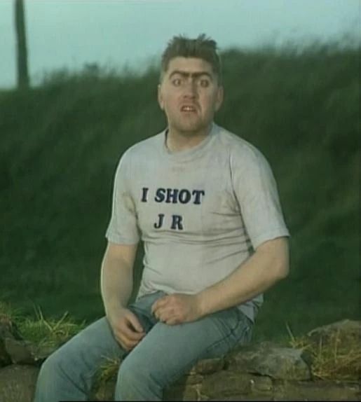 Tom from Father Ted sitting on a stone wall wearing a t-shirt that says: "I shot JR" 