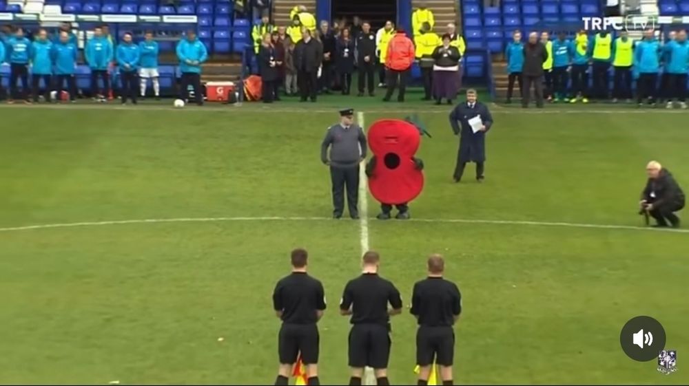 An military officer on a soccer field standing next to a poppy mascot 