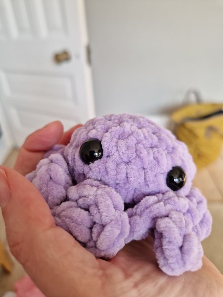 A purple crochet kraken with curly tentacles and big eyes is sitting in the palm of my hand 