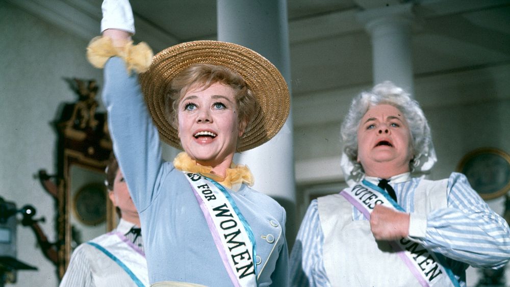 Screen of Marry Poppins with Mrs Banks and one of her maids  wearing sashes that read "Votes for Women," reminiscent of the suffragette movement. Mrs Banks in the foreground is smiling and raising her arm, wearing a light blue dress and a straw hat with yellow decorations. Another woman stands behind her, looking determined.