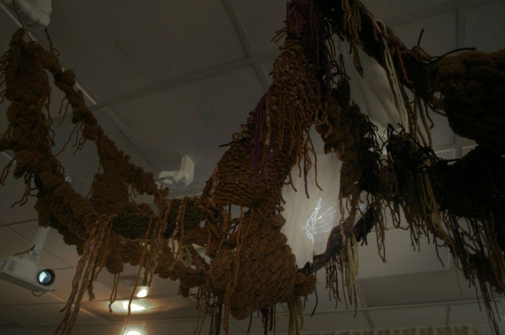 Yarn sculpture made of mostly brown yarn with some purple, beige, purple, and black strands. It is being held up by foam hands that are painted white. It is hanging from a ceiling. There is a small white mini projector. There is a white sheet in between the yarn with a projection of a peacock. 