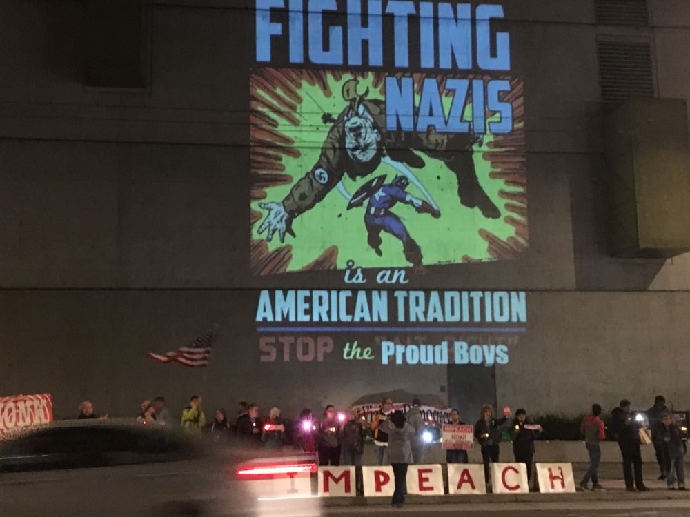 A group of citizens holding lights along Mission Street in San Francisco in 2019, with signs spelling out IMPEACH and a projection on the wall of the building depicting an illustration of someone sending a Nazi with a flying punch and the caption: FIGHTING NAIS is an American Tradition. STOP the Proud Boys.