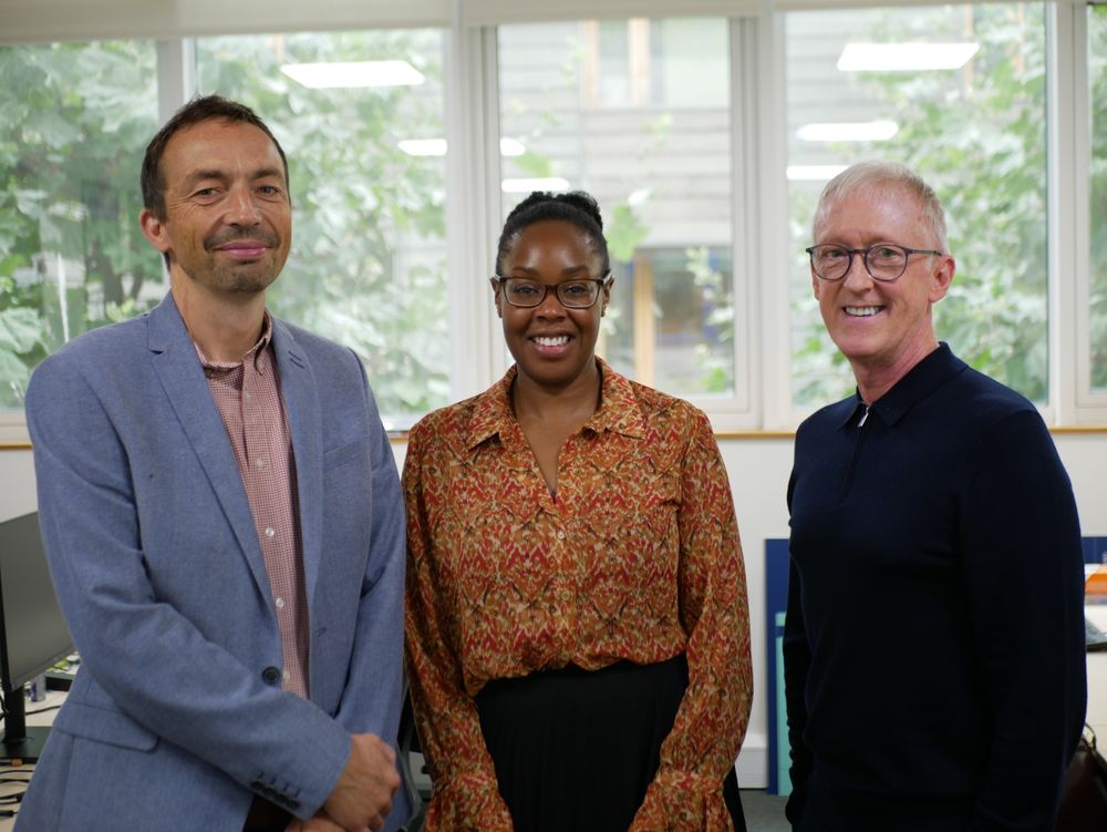 A group photo of our three new board members. All three are standing in front of a window and are smiling. Rick is wearing a pink shirt and a grey-blue jacket. Lara wears black glasses and a colourful red and orange shirt. Eddie wears glasses and a dark top.