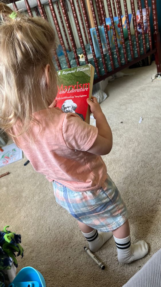 photo of a blond toddler with messy hair in a messy room covered with crayons holding a copy of Raymond Williams’ Marxism and Literature from ~2005. It has a bright red cover with a classic picture of bearded wise Karl Marx on it.