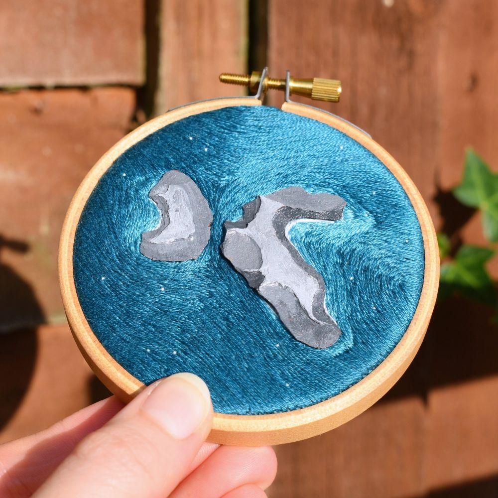 A hand holding a mini embroidery hoop, which depicts an aerial landscape consisting of blue flowing water around painted rocks.