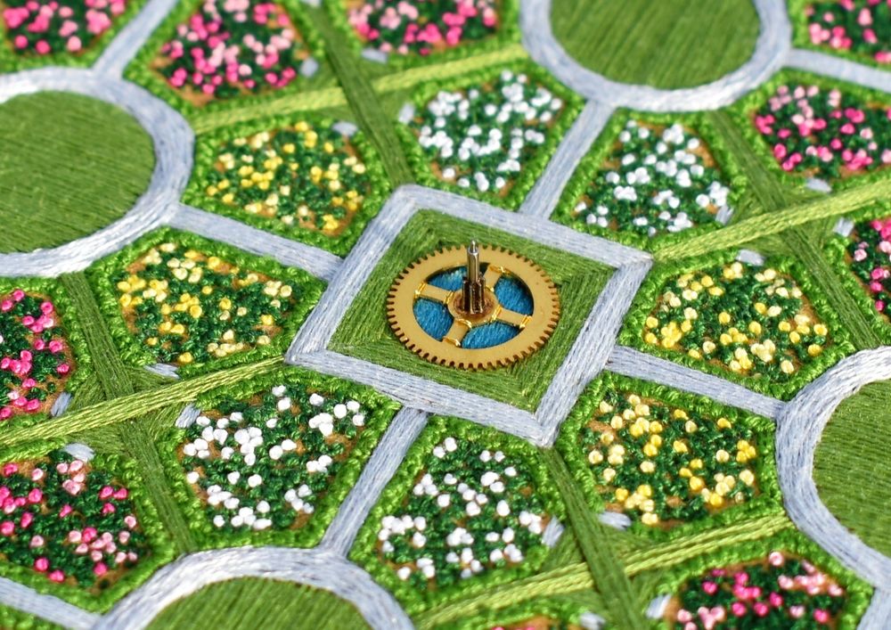 A tilted photo of an embroidered aerial-view landscape. It depicts a geometric rose garden with a fountain in the centre made from a repurposed clock cog.