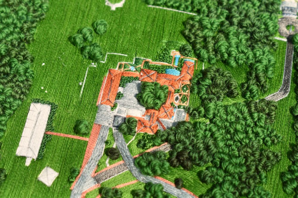 An aerial view embroidered landscape. It depicts red brick buildings and pathways surrounded by green lawn and trees, with a white tent to the left.