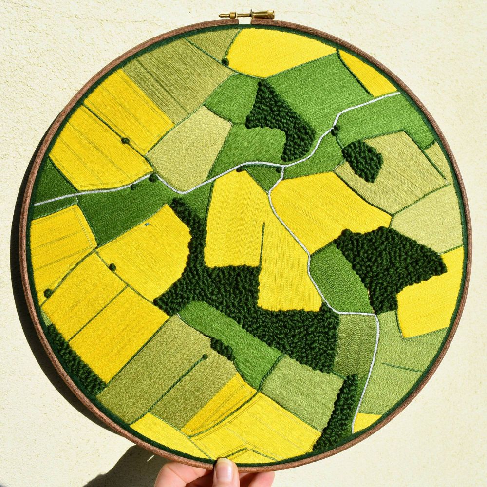 A hand holding an embroidery hoop in front of a cream wall. The hoop contains a sewn aerial landscape depicting oilseed meadows in bright yellow, green grass fields and areas of deep green trees. There's a grey road that breaks into two passing through the whole landscape.