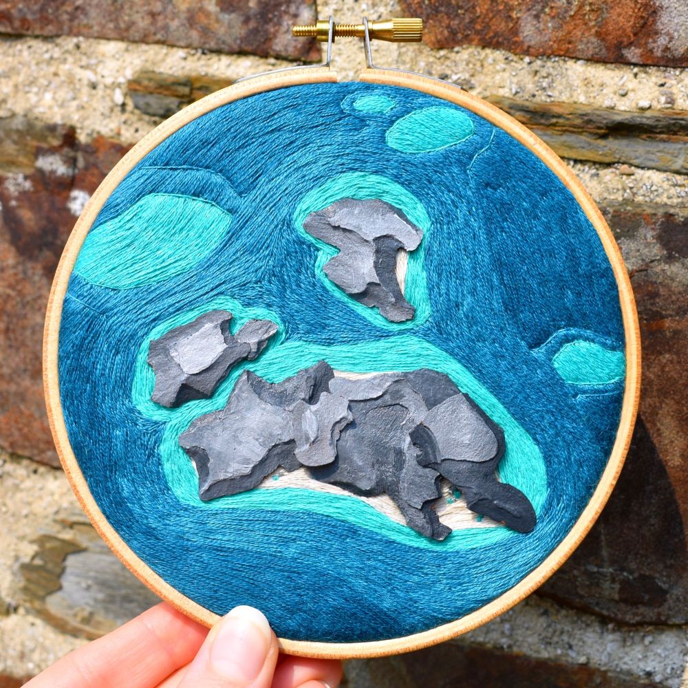 An embroidery hoop containing an embroidered aerial view landscape of the ocean. It contains rocky islands made from painted pine bark.