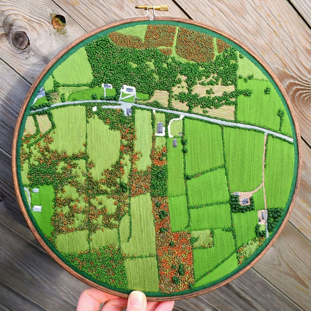 An embroidery hoop containing an embroidered aerial view landscape. It shows a road cutting through bright green meadows, brown rough land and houses.