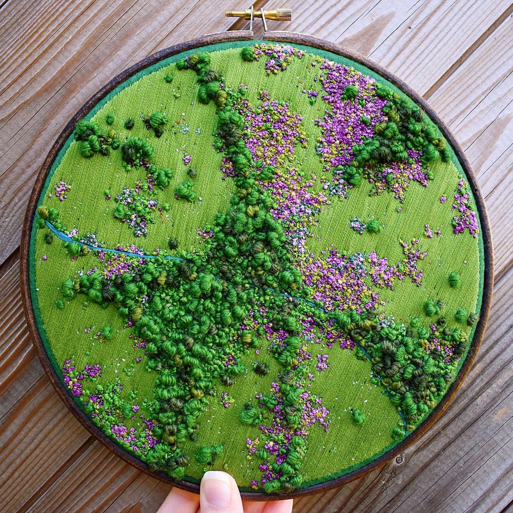 A hand holding an embroidery hoop against a wooden background - the hoop contains a sewn landscape depicting an aerial view of moorland. The majority is green trees and grass with large areas of heather bushes in a variety of purple shades. A tiny blue stream cuts across it all from W to SE.