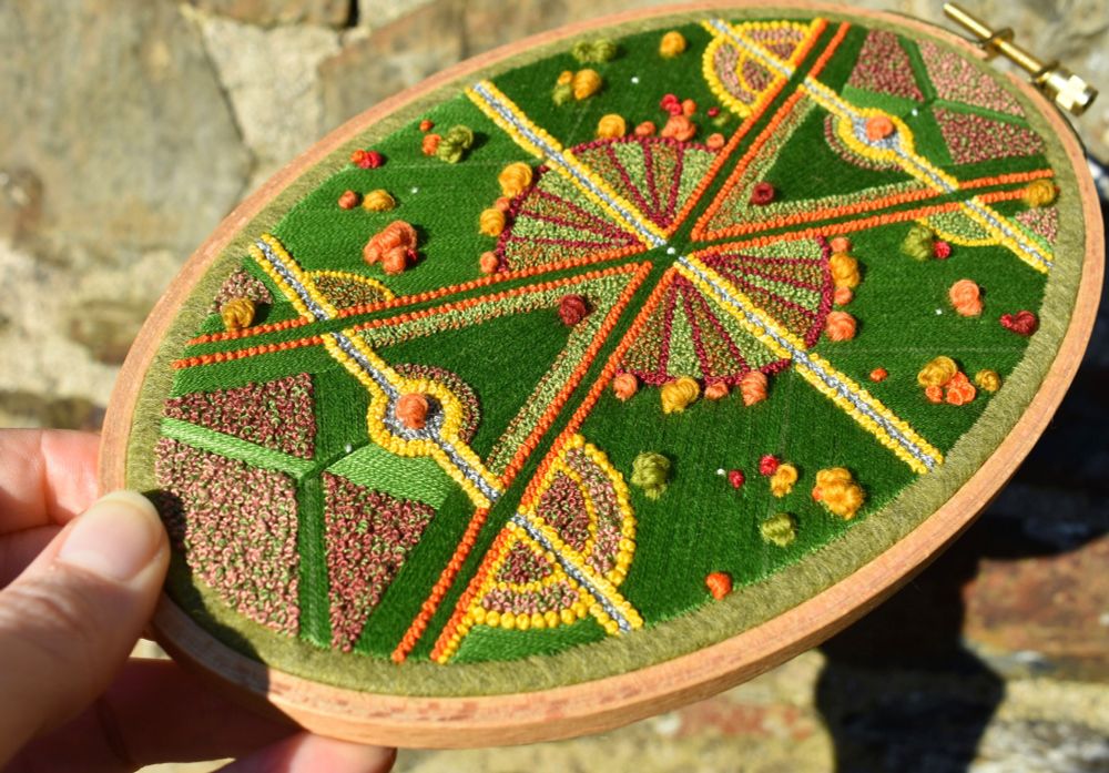 An embroidery hoop depicting an aerial formal garden. Autumn trees and bushes lines criss-crossing paths and blooming plant beds.