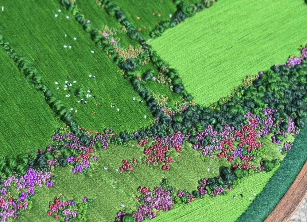 A close-up of a sewn aerial view landscape. It consists of bright green grass, knots trees and heather bushes in shades of purple.