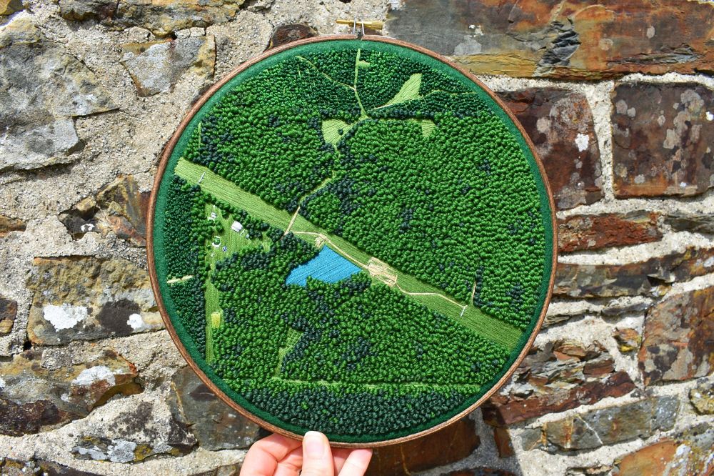 An embroidery hoop containing a sewn aerial view landscape. It mainly consists of French knot trees with a grass path cutting through and a central pond.