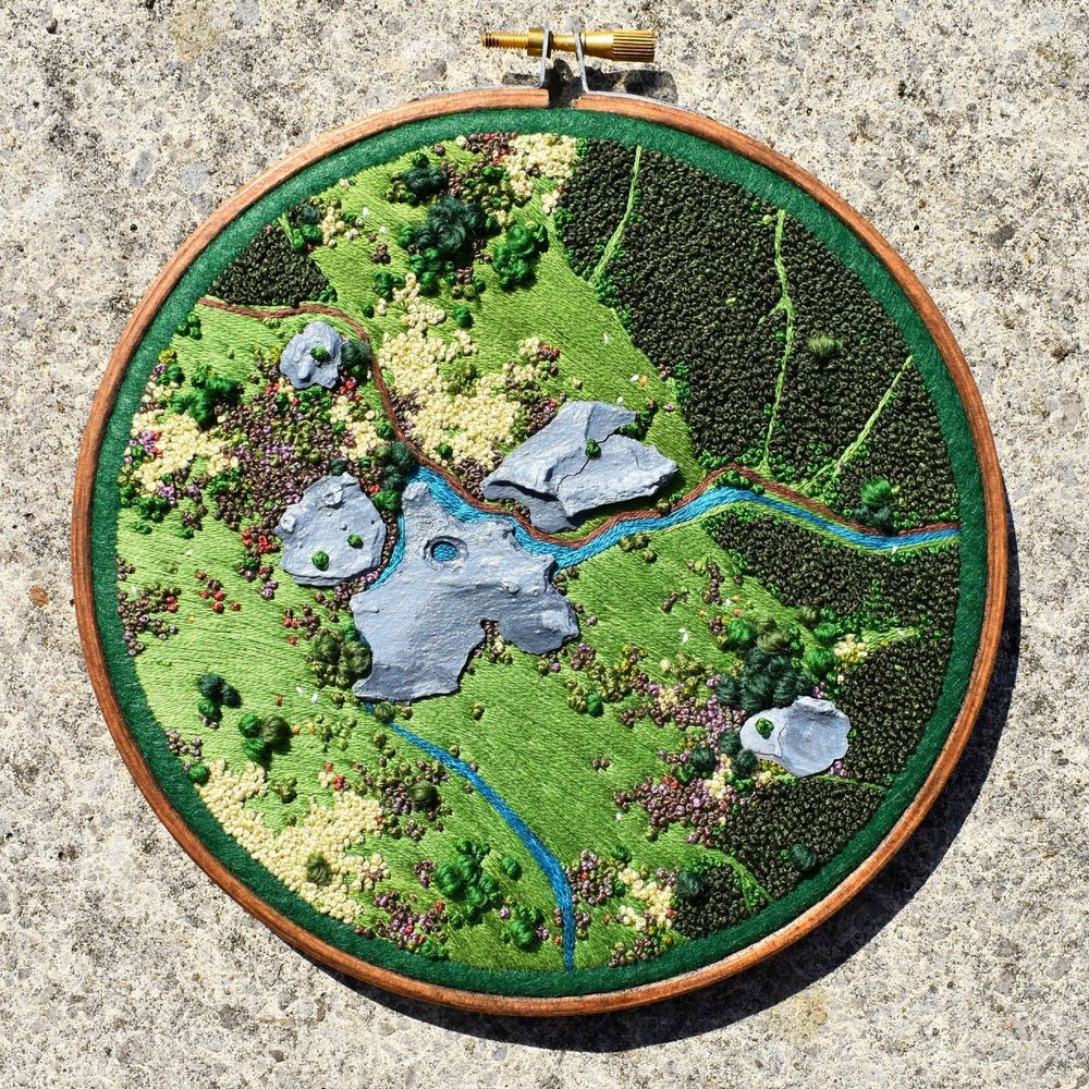 An embroidery landscape in a hoop frame - it depicts an embroidered aerial-view landscape of wild moorland, which includes a soft stream/brook, shrubs, scrubland and 'rocks' made from painted pine bark chips.