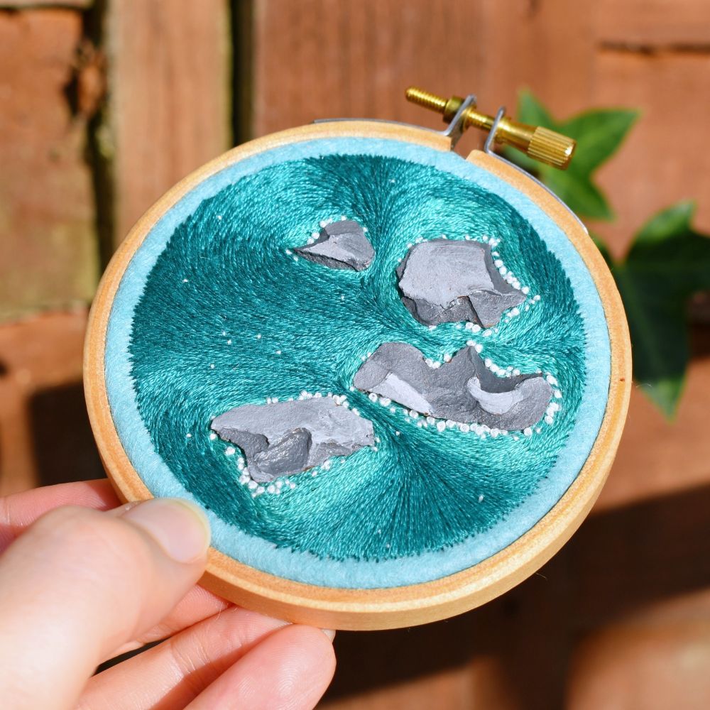 A hand holding a mini embroidery hoop, which depicts an aerial landscape consisting of blue flowing water around painted rocks.