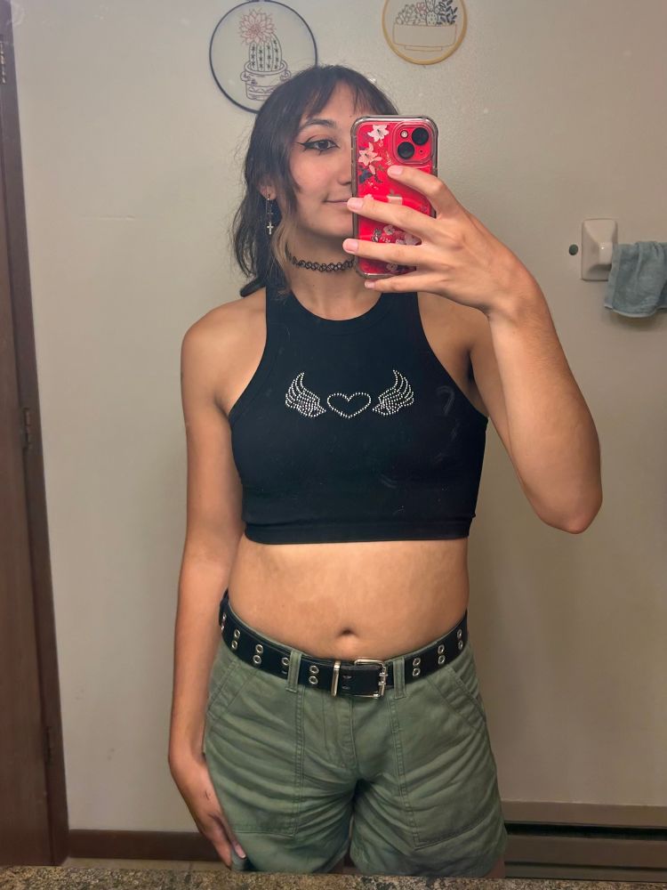 Me smiling in the mirror with the same eyeliner, green cargo shorts, and a small black tank top with a heart and wings on it