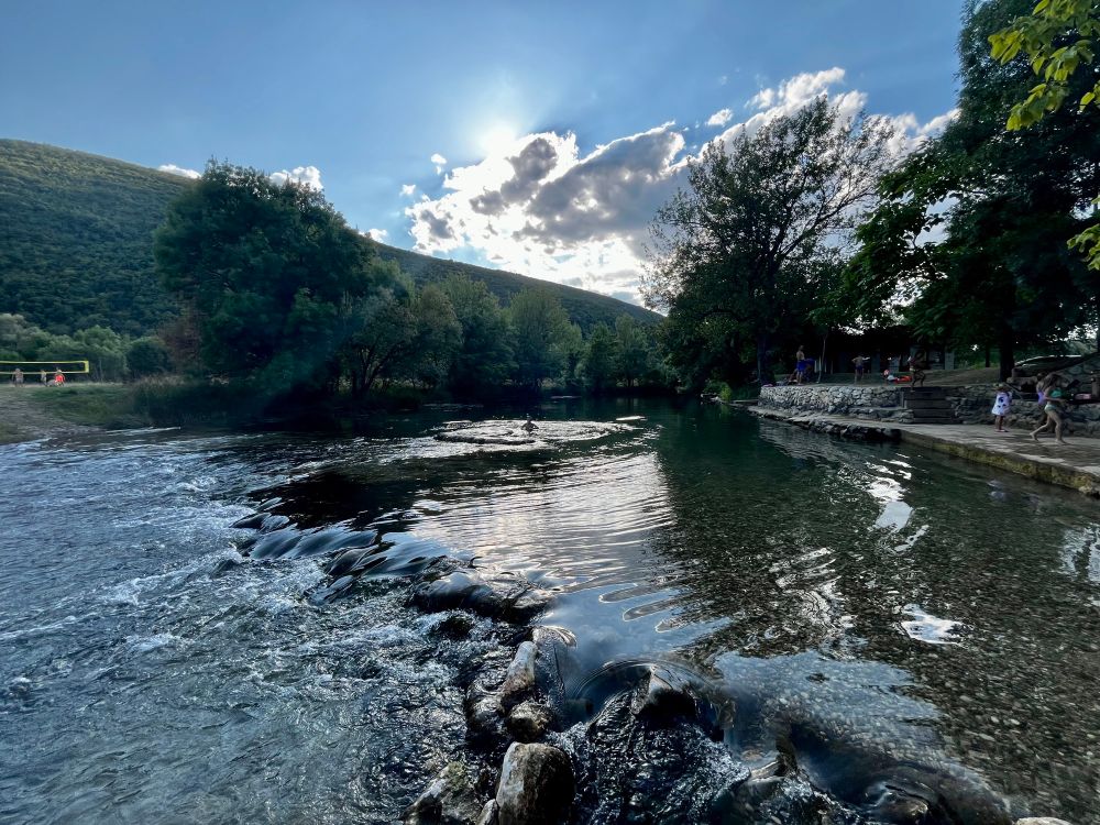 Fluss mit kleinem Wasserfall in der unteren Bildhälfte, darüber Bäume und ein blauer Himmeln mit ein paar Wolken, die die Sonne verdecken.
Rechts auf 3 Uhr erkennt man ein befestigtes Ufer, das als Badestelle dient.