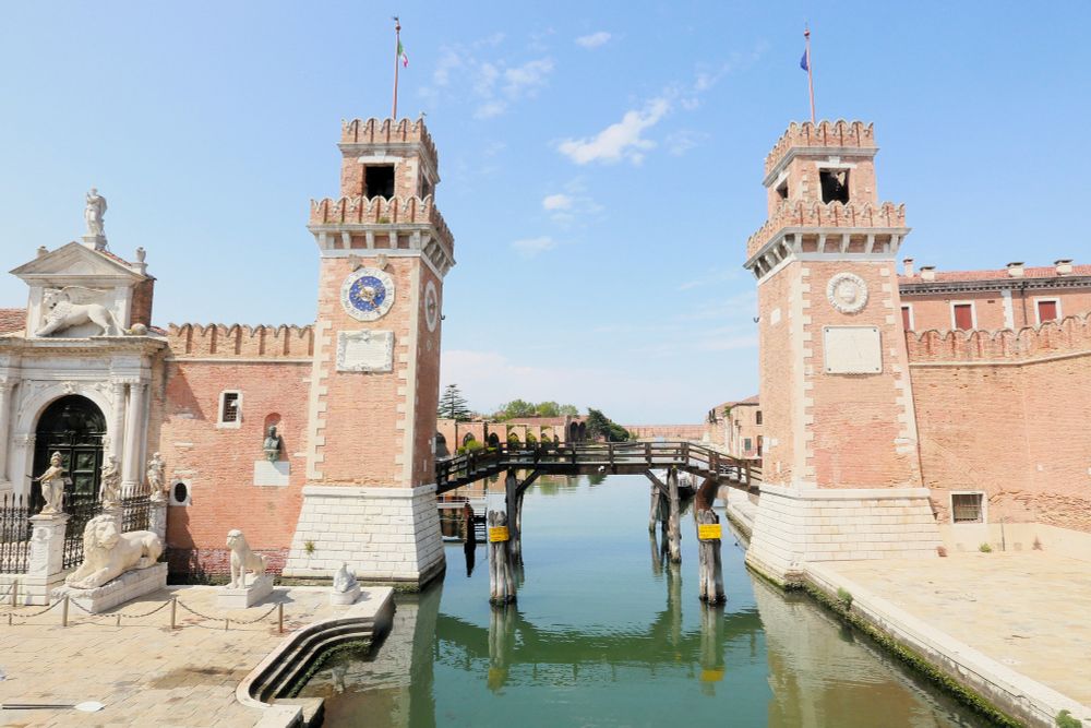Entrance to the Venice Arsenal. Two redbrick towers sit on opposite sides of a canal, with a wooden bridge crossing the canal behind them.