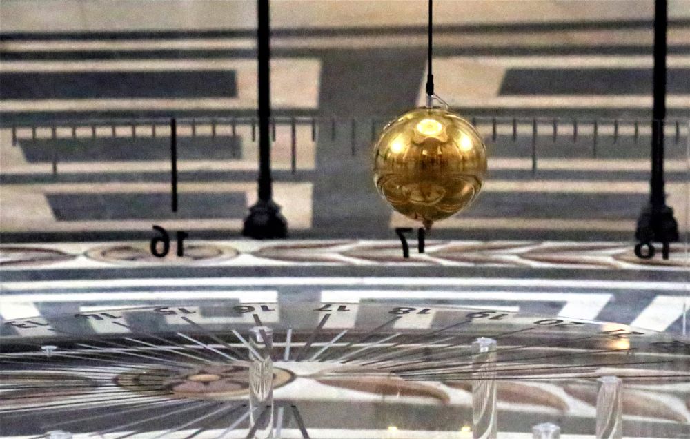 A long, golden pendulum swings over a marked out floor.