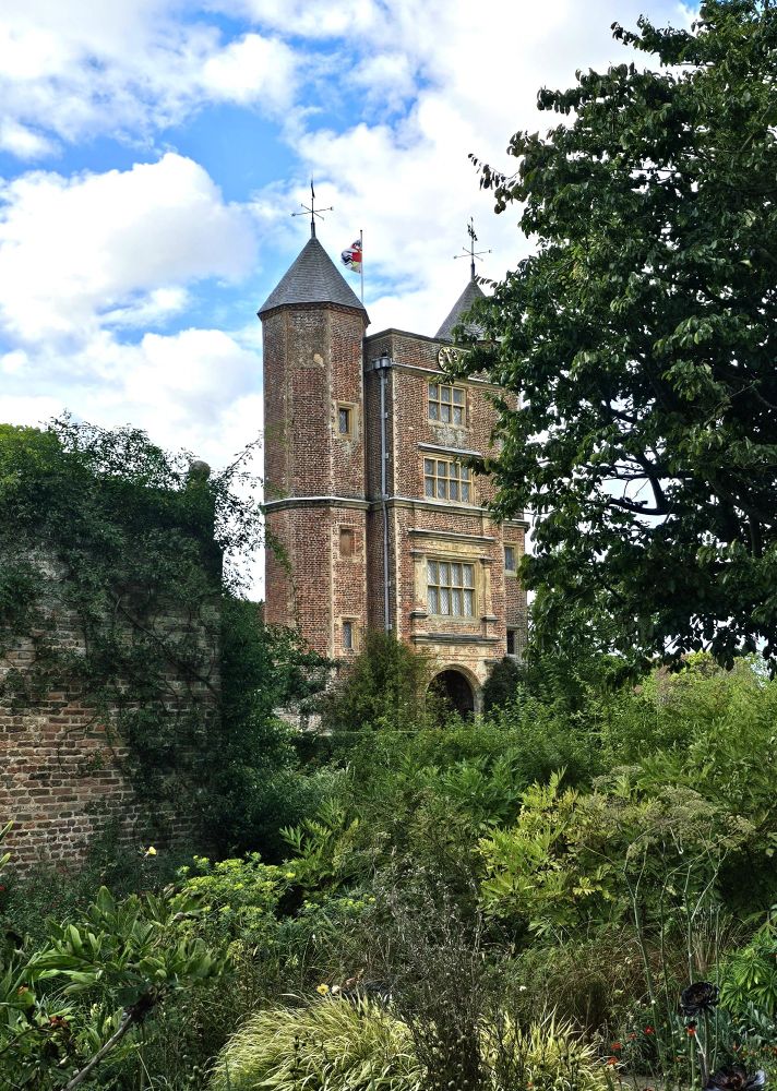 A tall, tudor gatehouse with two towers, in red brick, sits in the middle of a lush garden.