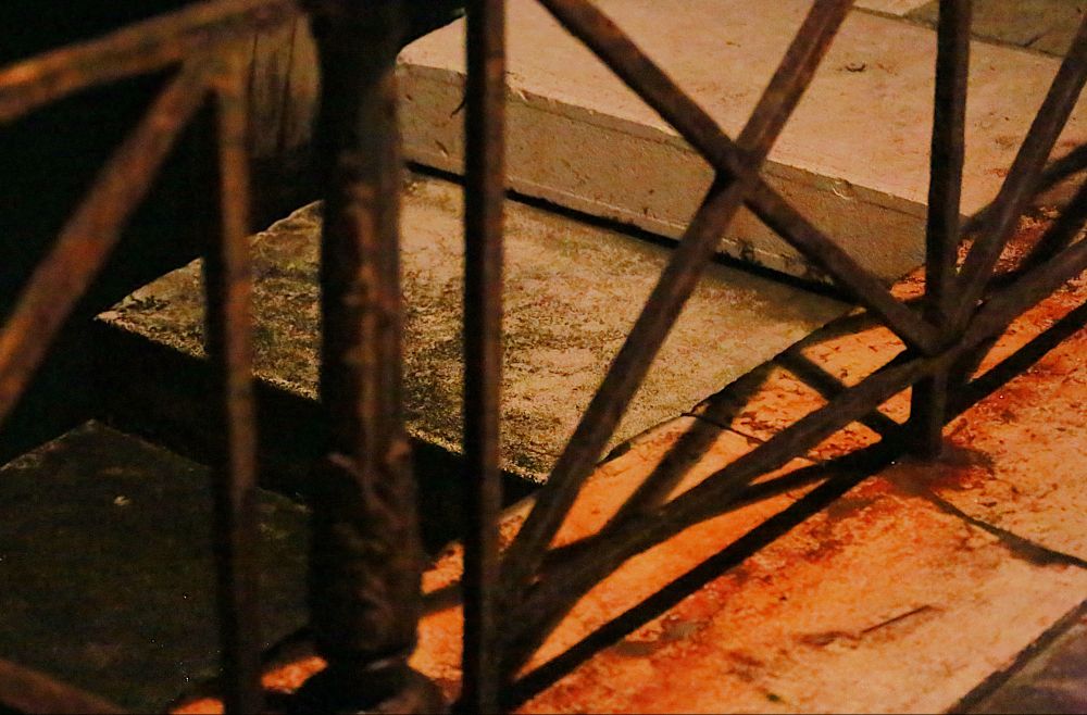 Stone steps leading down to water, with iron railings in the foreground. Below the railings are orange rust stains. The scene is shot at night, and there are strong shadows.