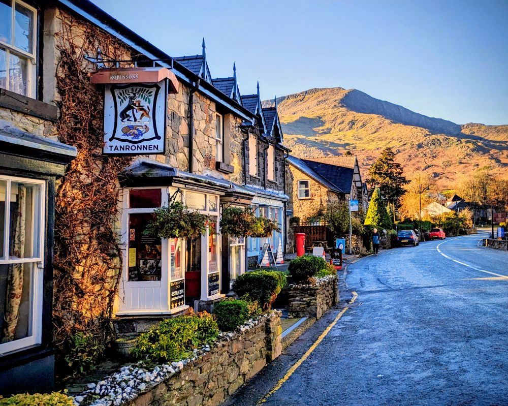 A quaint, rustic Welsh inn with a stone facade and climbing ivy, named "Tanronnen," stands proudly in Beddgelert village.

The inn's sign features the tragic legend of a faithful dog named Gelert who was mistakenly killed by his master, Prince Llywelyn the Great.

The warm glow of the rising sun illuminates the scene, highlighting the detailed architecture and the vibrant greenery around the building. A winding road stretches into the distance, with a few cars parked along the side, and a person walking on the pavement.

Majestic mountains rise in the background, their peaks bathed in the soft, golden light of dawn. The sky above is a clear, serene blue, adding to the picturesque charm of the rural gogledd Cymru landscape.